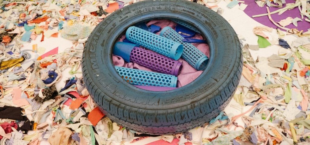 Su un pavimento ricoperto da ritagli di carta e stoffa è presente uno pneumatico blu con dentro oggetti blu e viola