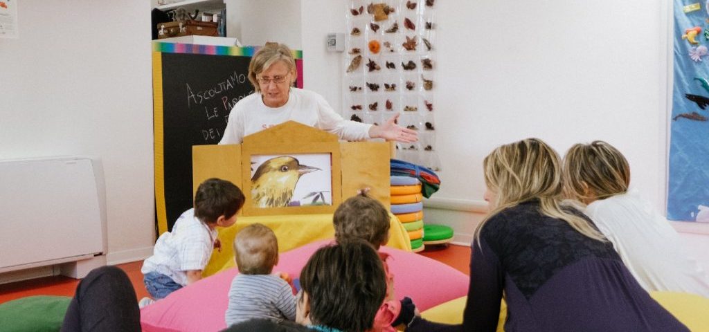 Un' insegnante mostra ai bambini in una cornice fatta a casetta il disegno di un uccello giallo. I bambini sono seduti con degli adulti su grossi cuscini