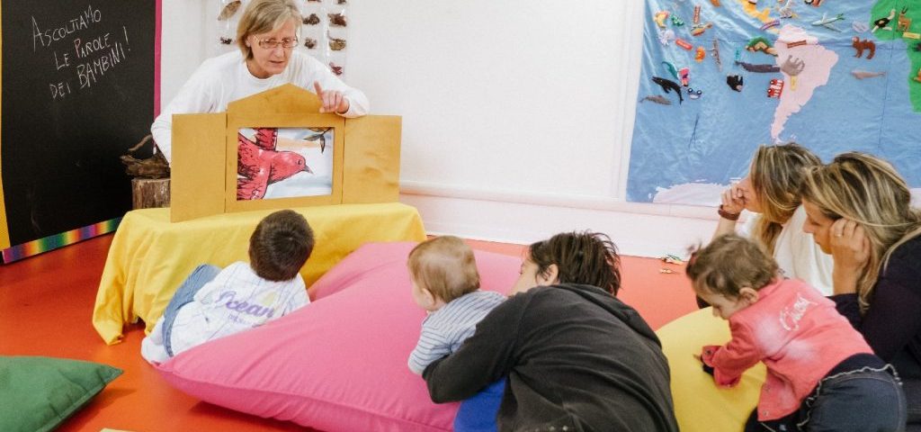 Un' insegnante mostra ai bambini in una cornice fatta a casetta il disegno di un uccello rosso. I bambini sono seduti con degli adulti su grossi cuscini