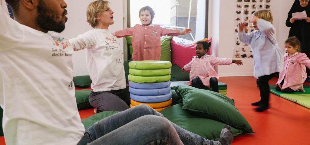 Bambini e insegnanti nella stanzaq di Spazio ZeroSei con il pavimento rosso muovono le braccia