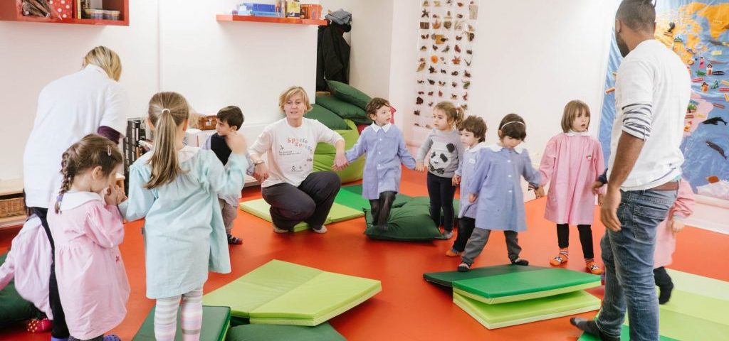 Tre insegnanti e alcuni bambini si tengono per mano formando un semicerchio nella stanza di Spazio ZeroSei con il pavimento rosso