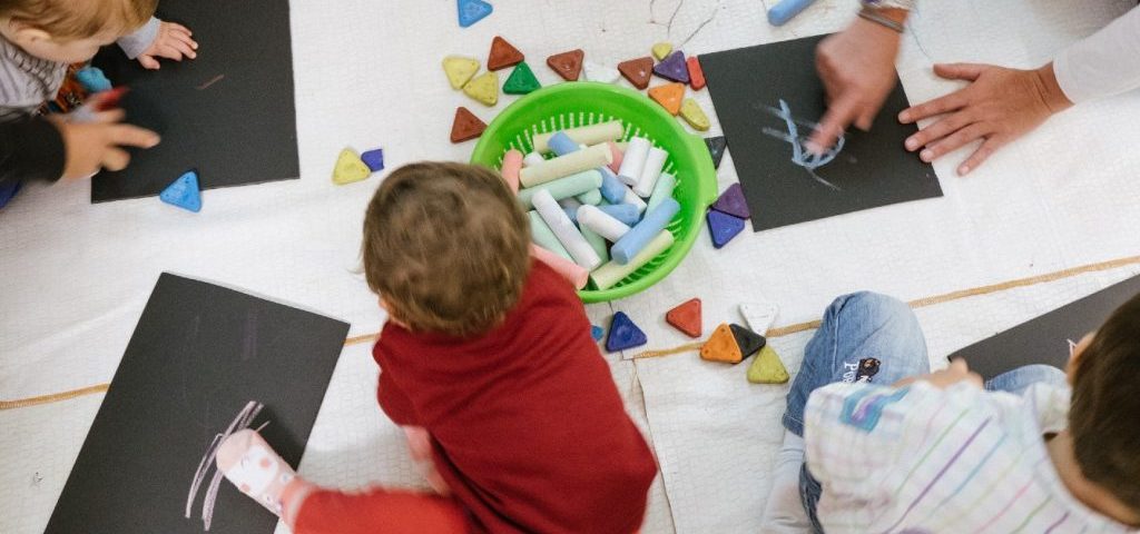 Dei bambini seduti a terra stanno colorando con dei gessetti su dei fogli neri con l'aiuto di due insegnanti