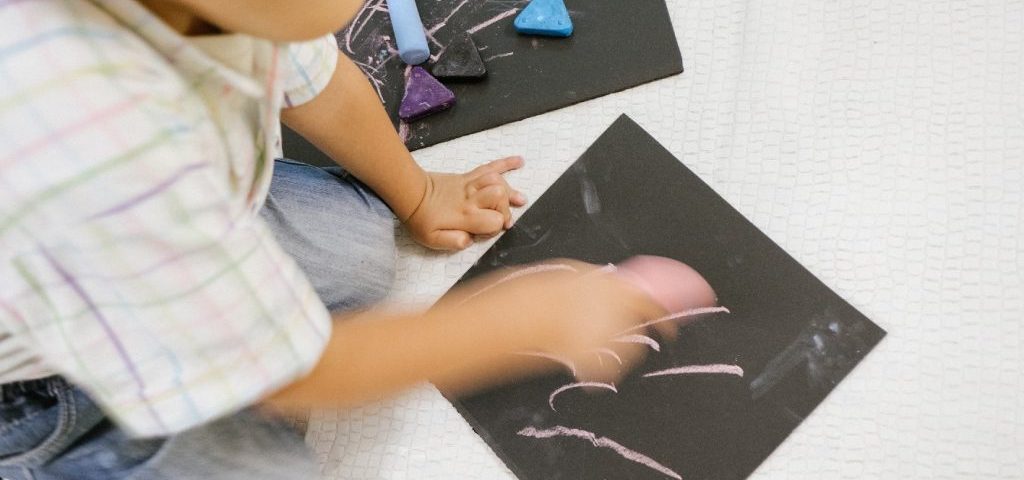 Un bambino seduto a terra sta colorando con dei gessetti su dei fogli neri