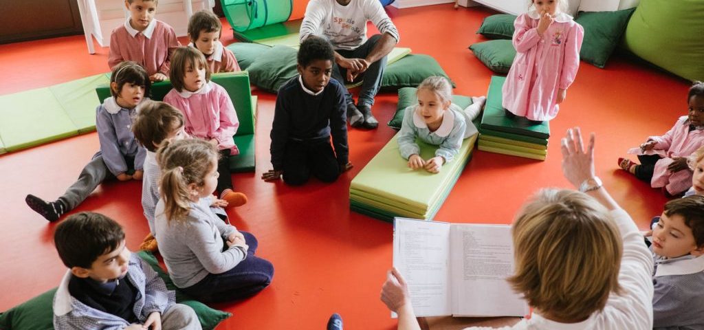 Un'insegnante sta leggendo un libro a un gruppo di bambini seduti a terra nella stanza di Spazio ZeroSei con il pavimento rosso.