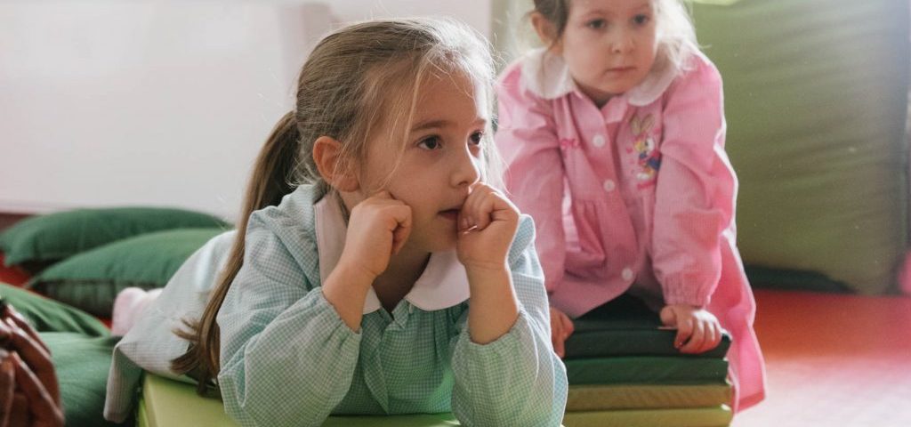 Due bambine, una seduta e una sdraiata, su dei materassini verdi nella stanza con il pavimento rosso