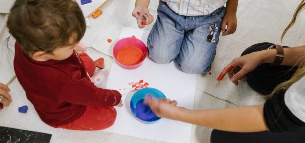 Due bambini visti dall'alto dipingono con le mani un grosso foglio bianco aiutati da un'insegnante