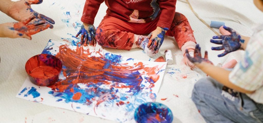 Due bambini aiutati da un'insegnante dipingono con le mani un grosso foglio bianco