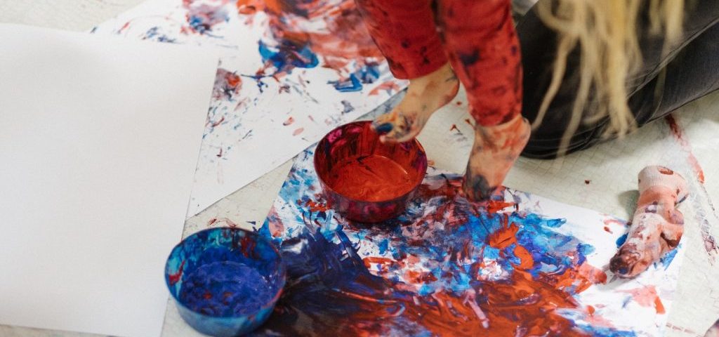 Un bambino sta dipingendo dei fogli bianchi immergendo i piedini in ciotole con tinte rosse e blu
