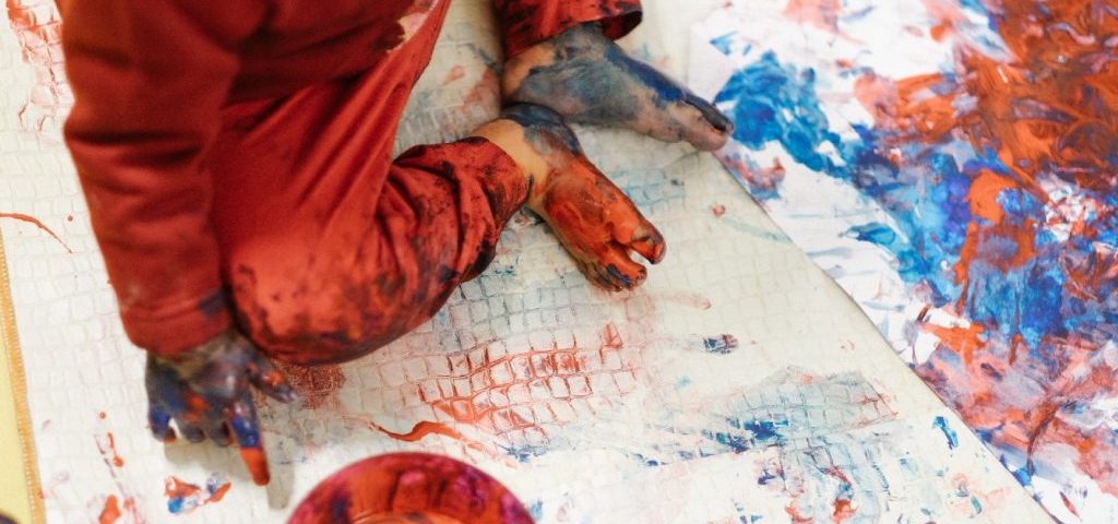 Un bambino tutto sporco di pittura (mani, piedi, vestiti) sta seduto su un grosso telo dipinto di rosso e di blu
