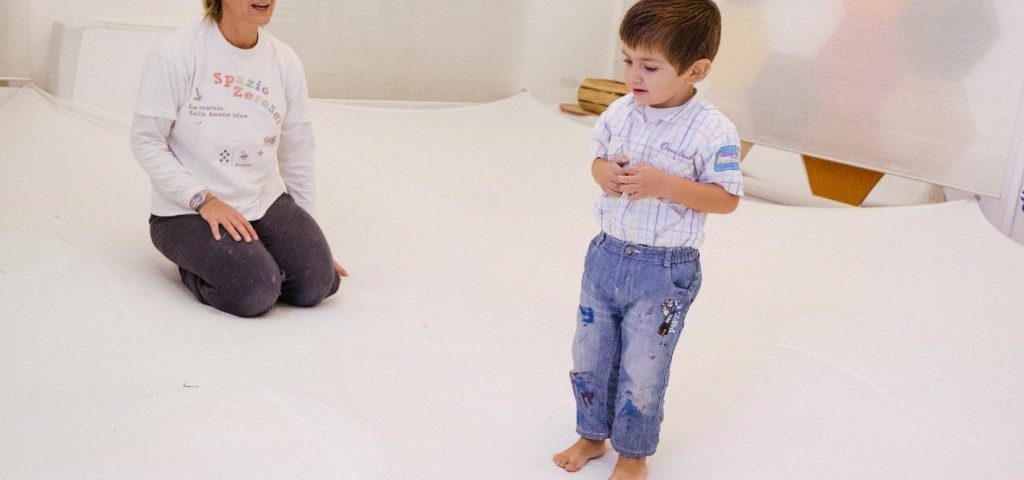 Un bambino gioca con un'insegnante in una stanza con pareti bianche e pavimento ricoperto da un grosso telo bianco.