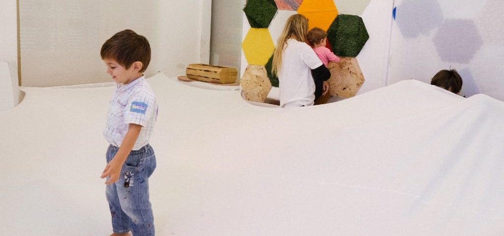 Dei bambini giocano in una stanza con pareti bianche e pavimento ricoperto da un grosso telo bianco. Su una parete sono dipinti esagoni colorati