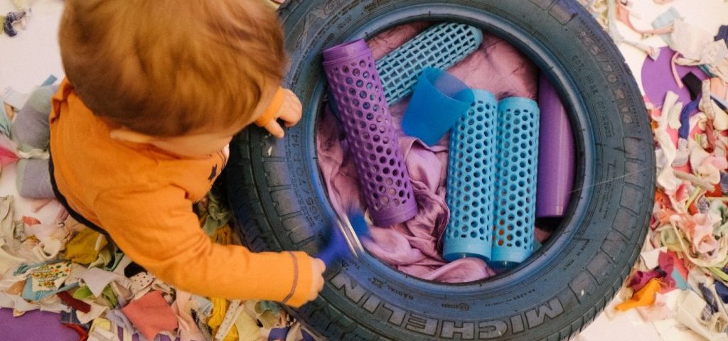 Su un pavimento ricoperto da ritagli di carta e stoffa, un bambino gioca con degli oggetti colorati messi dentreo uno pneumatico