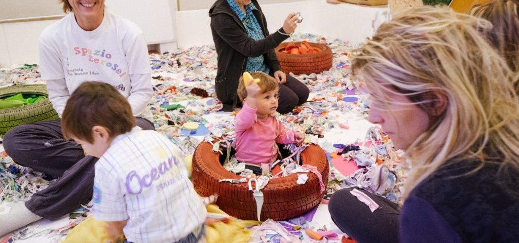 Su un pavimento ricoperto da ritagli di carta e stoffa, due bambini giocano con insegnanti e genitori. Una bambina è seduta in uno pneumatico dipinto di arancione