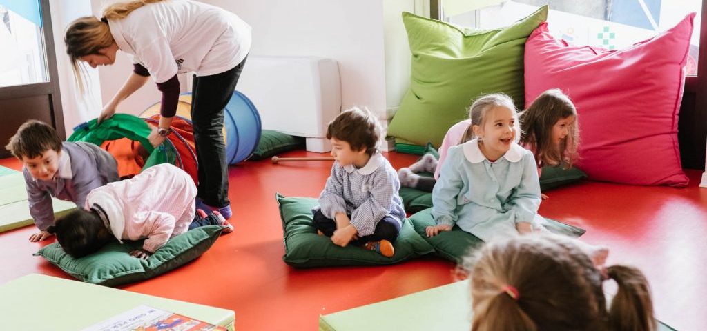 Nella stanza con il pavimento rosso sono presenti un tunnel da gioco, grossi cuscini e materassini colorati. Un gruppo di bambini è seduto a terra su cuscini verdi