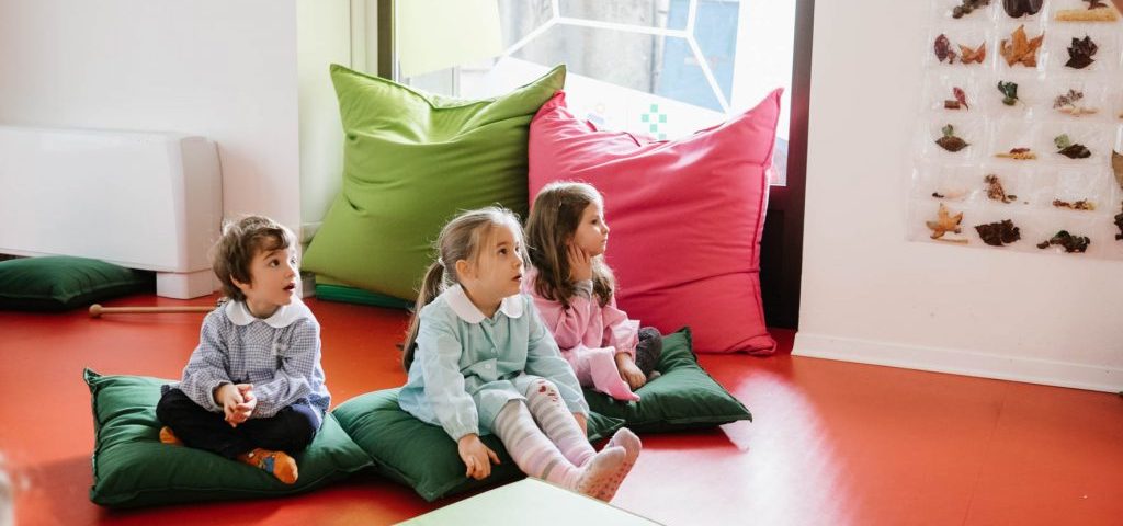 Alcuni bambini seduti a terra su grossi cuscini osservano meravigliati una scena in alto