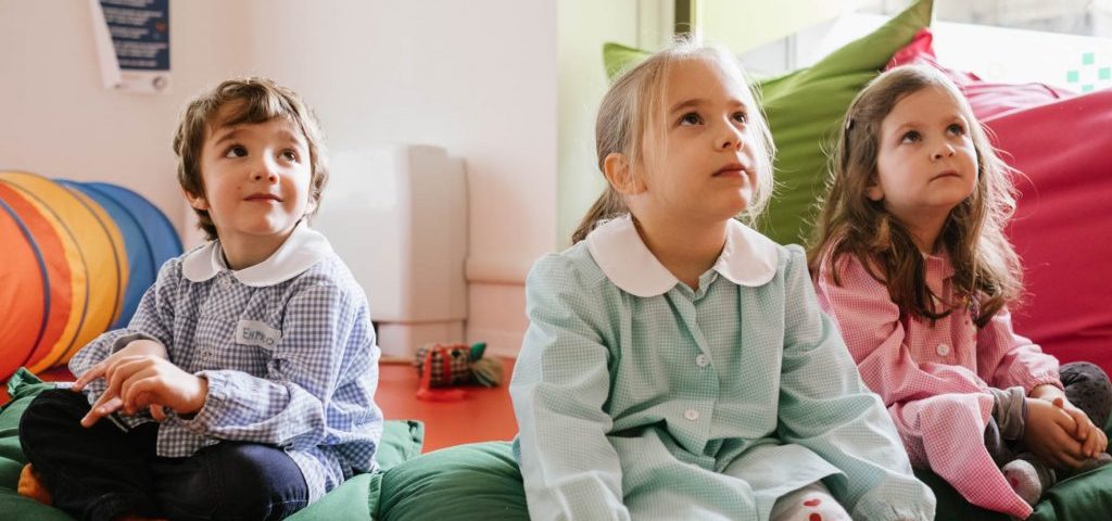 Alcuni bambini seduti a terra su grossi cuscini osservano meravigliati una scena in alto