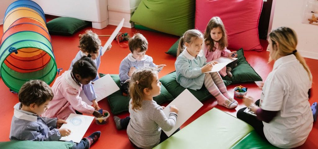 Nella stanza con il pavimento rosso sono presenti un tunnel da gioco, grossi cuscini e materassini colorati. Un gruppo di bambini sta disegnando su fogli bianchi aiutati da un'insegnante.