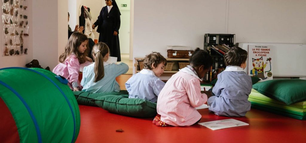 Un gruppo di bambini seduti su un pavimento rosso in una stanza di Spazio ZeroSei. Alcuni bambini sono seduti su cuscini verdi, mentre altri sono sul pavimento.