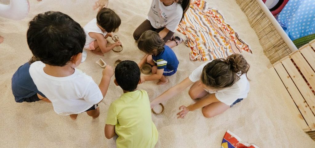 Un gruppo di bambini gioca nella sabbia con un'insegnante nella sala allestita per l'estate